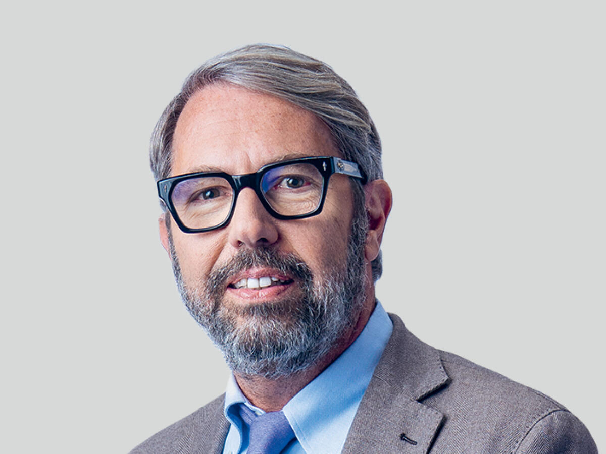 A professional portrait of a middle-aged man with short, graying hair and a well-groomed beard, wearing black-framed glasses, a light brown suit, a light blue dress shirt, and a matching blue tie. He has a confident and approachable expression with a slight smile. The background is a neutral light gray gradient.