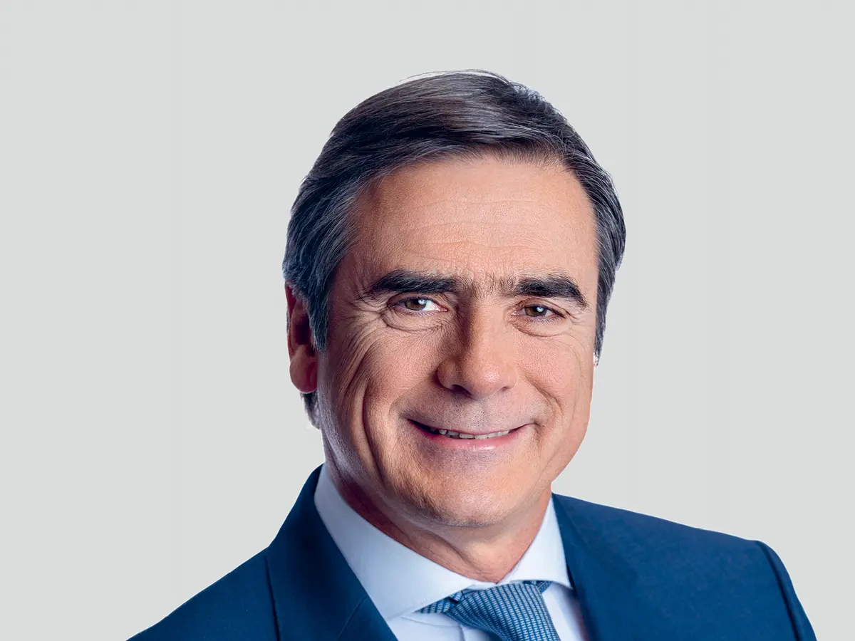 A professional portrait of a middle-aged man with short, dark gray hair, wearing a navy blue suit, a light blue dress shirt, and a matching blue tie. He has a confident and friendly expression with a slight smile. The background is a neutral light gray gradient.