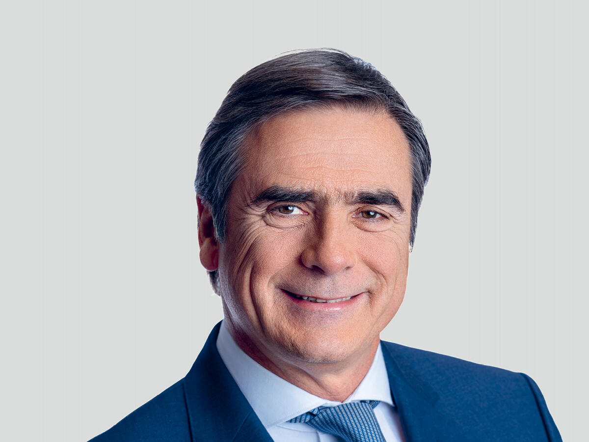 A professional portrait of a middle-aged man with short, dark gray hair, wearing a navy blue suit, a light blue dress shirt, and a matching blue tie. He has a confident and friendly expression with a slight smile. The background is a neutral light gray gradient.