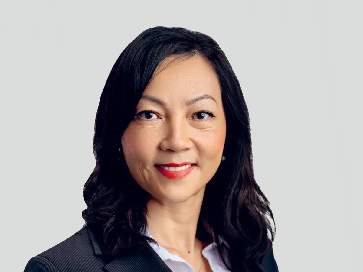 A professional portrait of a middle-aged woman with long, wavy black hair, wearing a black blazer over a white blouse. She has a confident and friendly expression with a slight smile and red lipstick. The background is a neutral light gray gradient.