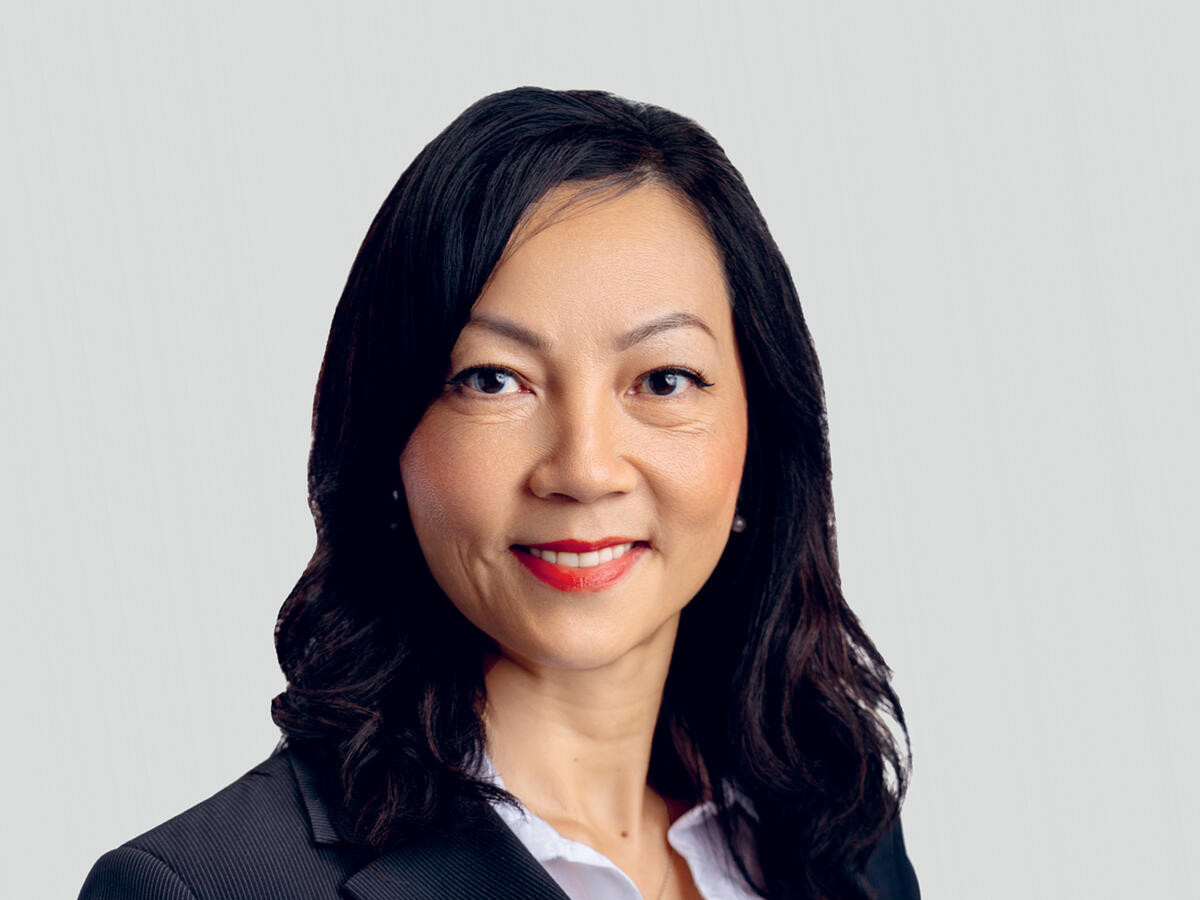A professional portrait of a middle-aged woman with long, wavy black hair, wearing a black blazer over a white blouse. She has a confident and friendly expression with a slight smile and red lipstick. The background is a neutral light gray gradient.