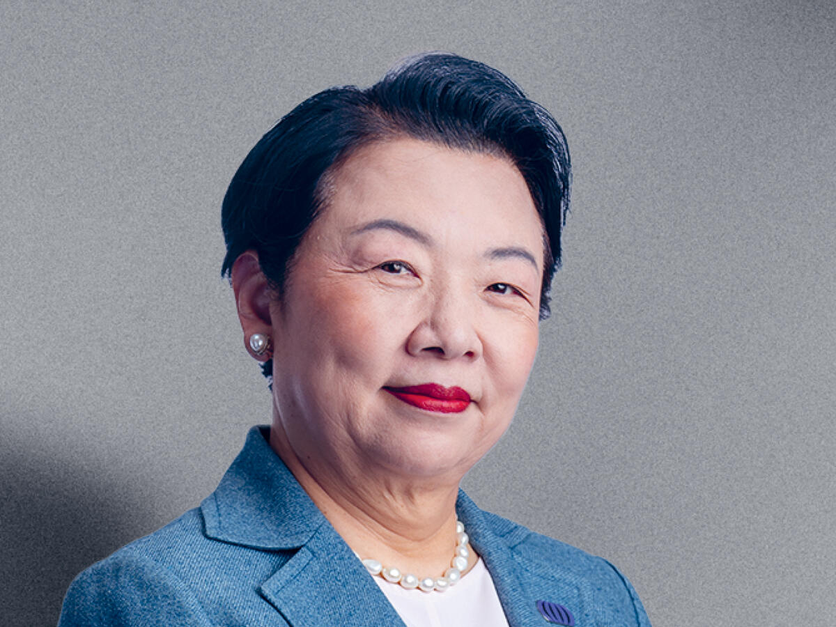 A professional portrait of a middle-aged woman with short black hair, wearing a light blue blazer over a white blouse, accessorized with pearl earrings and a pearl necklace. She has a confident and poised expression with a slight smile. The background is a neutral gray gradient.