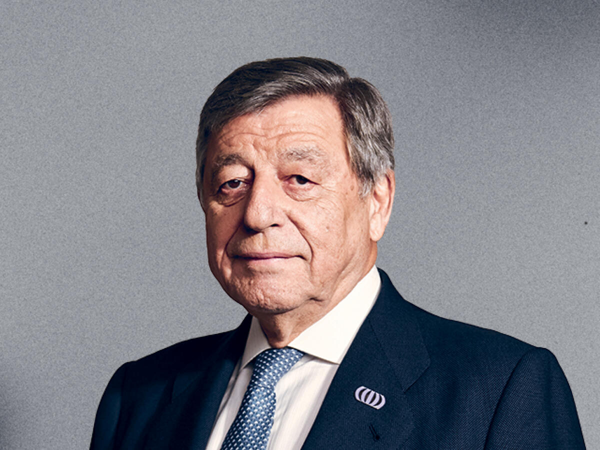 A professional portrait of an older man with short gray hair, wearing a dark navy suit, a white dress shirt, and a blue patterned tie. He has a serious yet approachable expression with a slight smile. A lapel pin is visible on his suit jacket. The background is a neutral gray gradient.