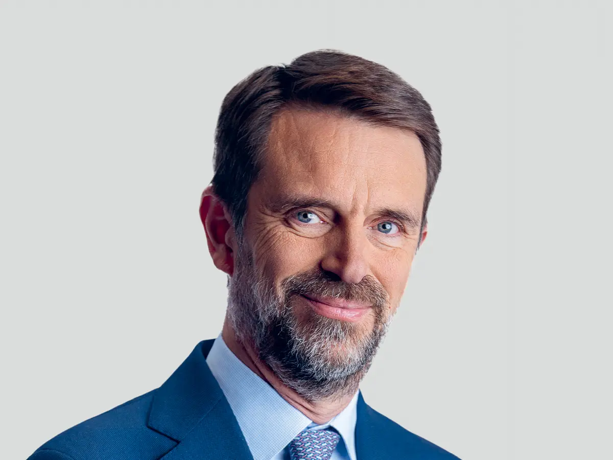 A professional portrait of a middle-aged man with short brown hair and a well-groomed beard, wearing a navy blue suit, a light blue dress shirt, and a patterned blue tie. He has a confident and approachable expression with a slight smile. The background is a neutral light gray gradient.