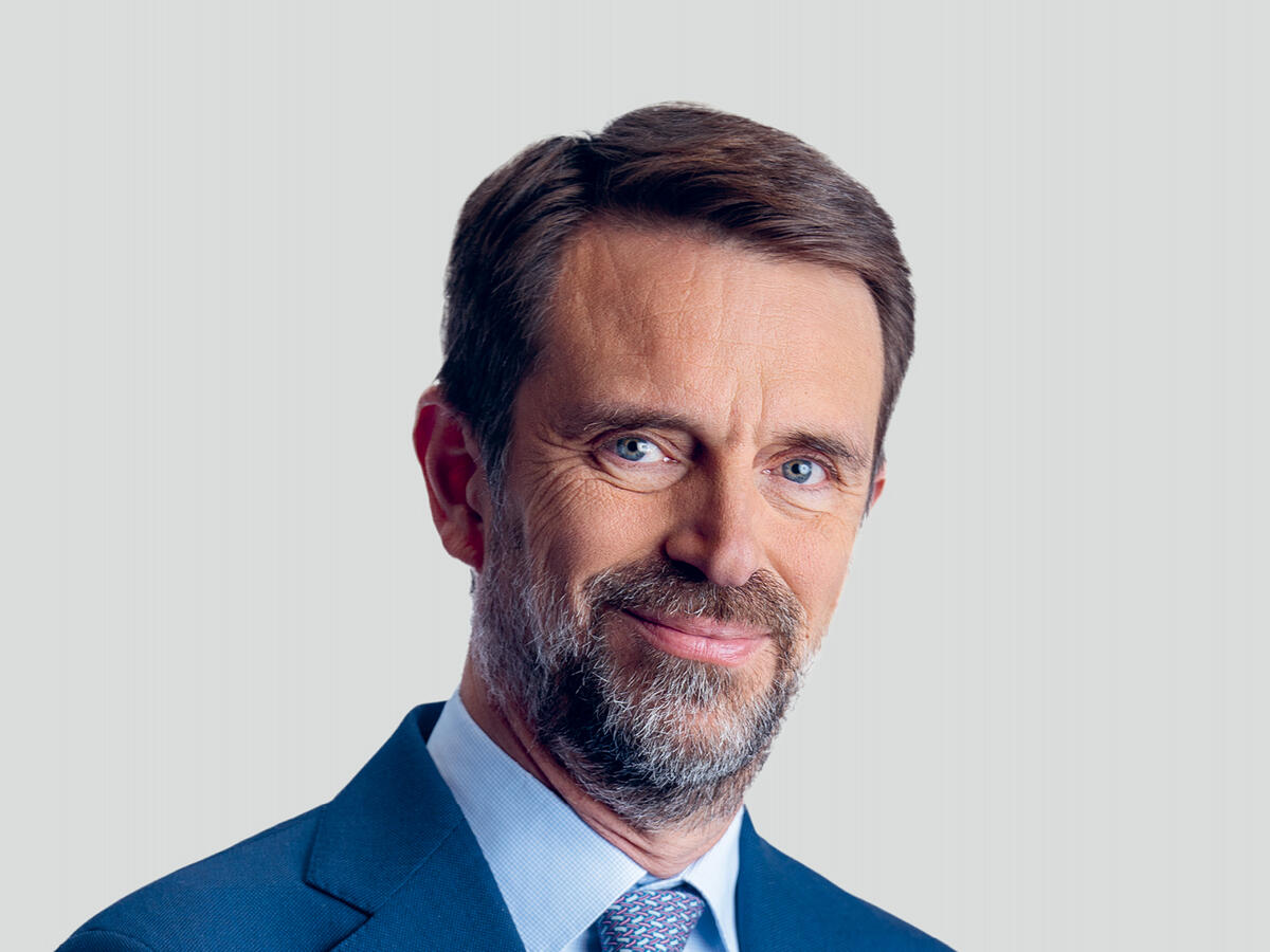 A professional portrait of a middle-aged man with short brown hair and a well-groomed beard, wearing a navy blue suit, a light blue dress shirt, and a patterned blue tie. He has a confident and approachable expression with a slight smile. The background is a neutral light gray gradient.