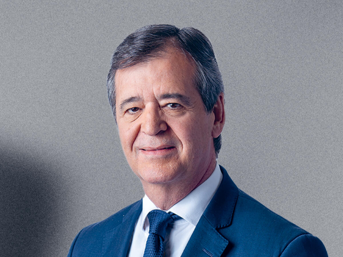 A professional portrait of a middle-aged man with short, dark gray hair, wearing a navy blue suit, a white dress shirt, and a dark blue tie. He has a calm and confident expression with a slight smile. The background is a neutral gray gradient.