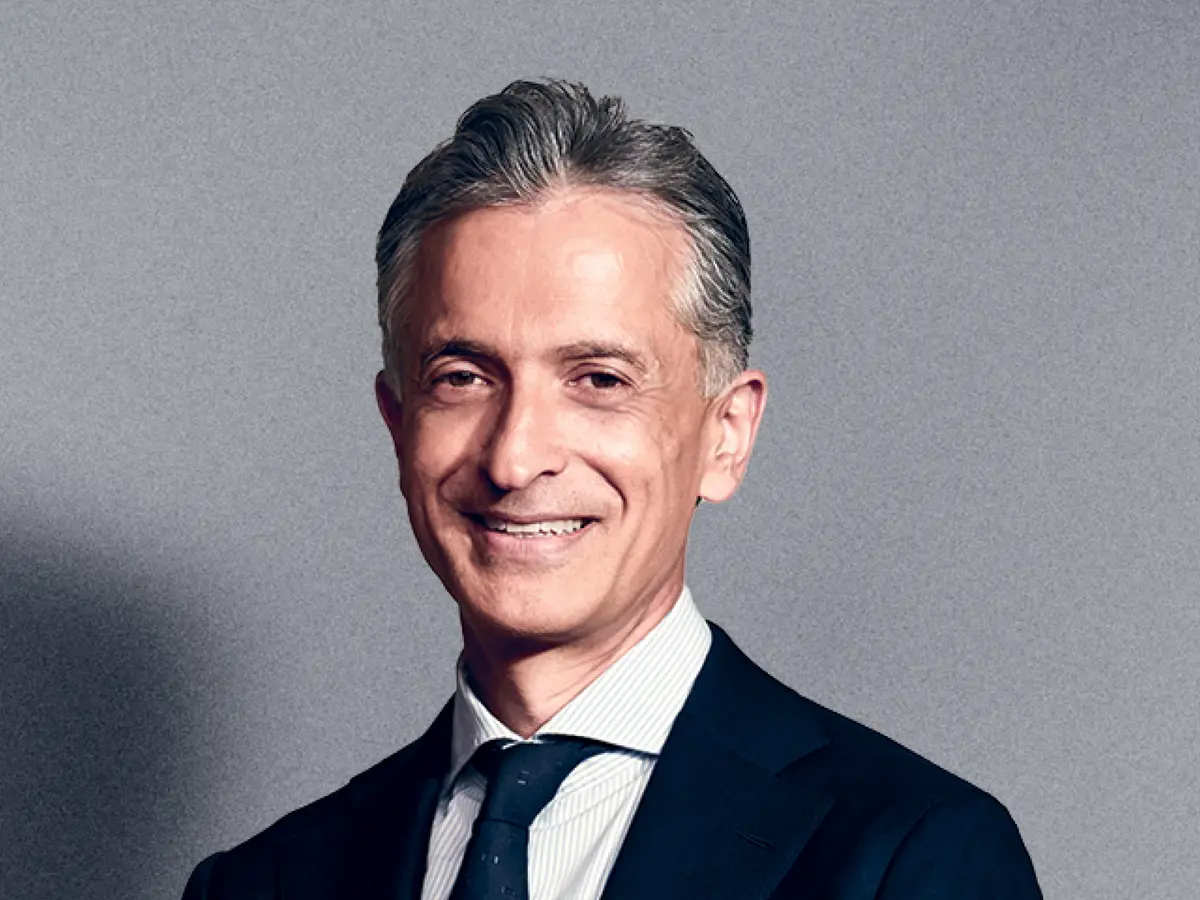A professional portrait of a middle-aged man with short, neatly styled salt-and-pepper hair, wearing a dark navy suit, a white dress shirt, and a dark tie. He has a warm and confident smile. The background is a neutral gray gradient.