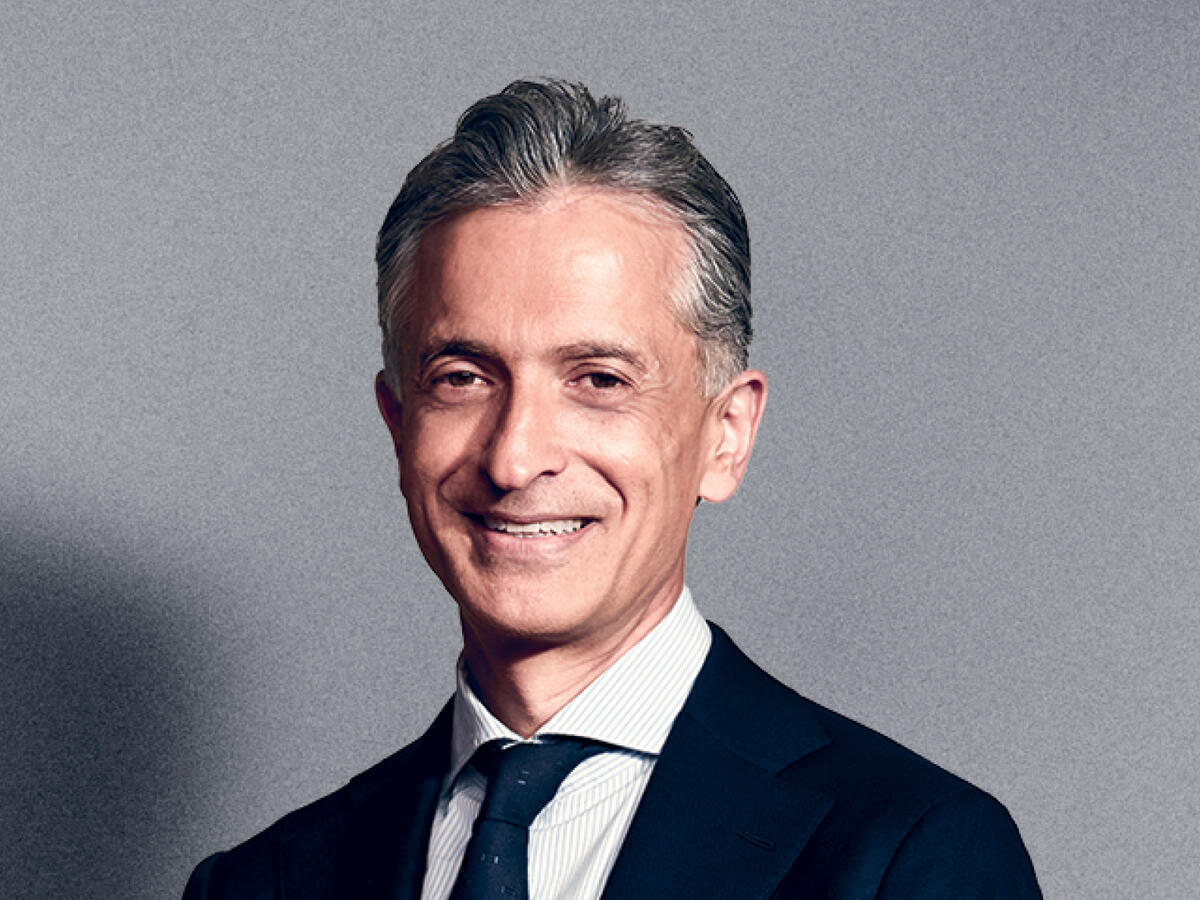 A professional portrait of a middle-aged man with short, neatly styled salt-and-pepper hair, wearing a dark navy suit, a white dress shirt, and a dark tie. He has a warm and confident smile. The background is a neutral gray gradient.