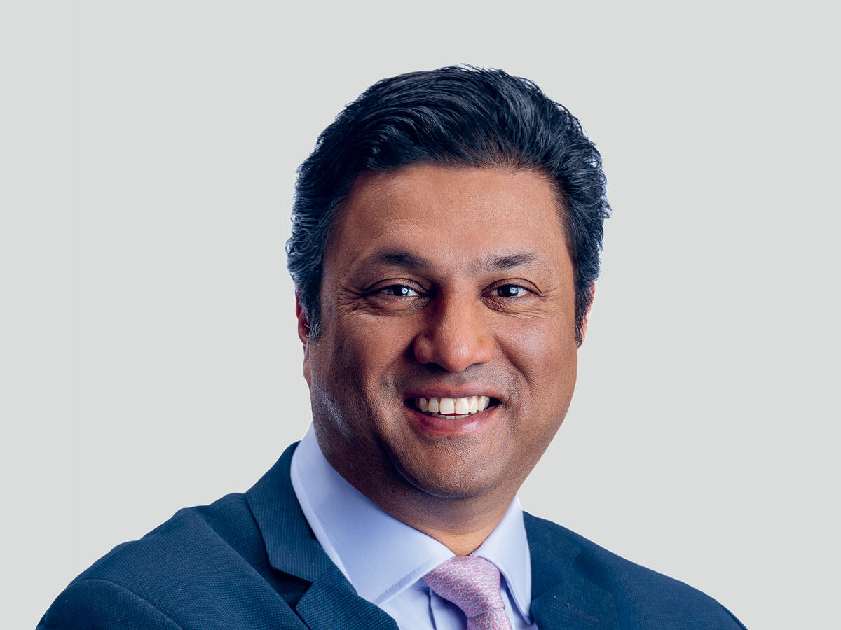 A professional portrait of a middle-aged man with short, wavy black hair, wearing a navy blue suit, a light blue dress shirt, and a pink patterned tie. He has a warm and confident expression with a broad smile. The background is a neutral light gray gradient.