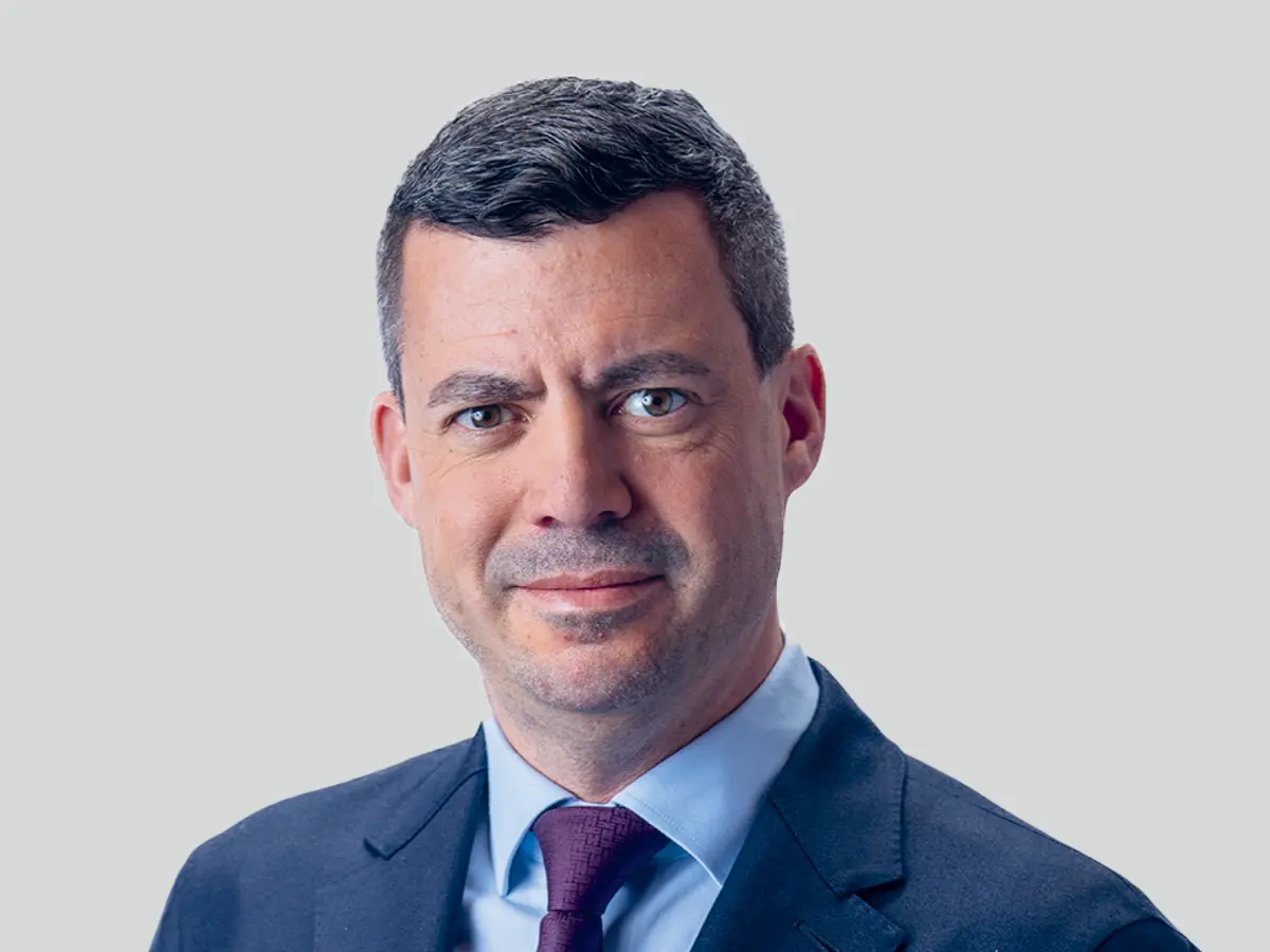 A professional portrait of a middle-aged man with short, dark gray hair, wearing a navy blue suit, a light blue dress shirt, and a dark purple tie. He has a confident and composed expression with a slight smile. The background is a neutral light gray gradient.