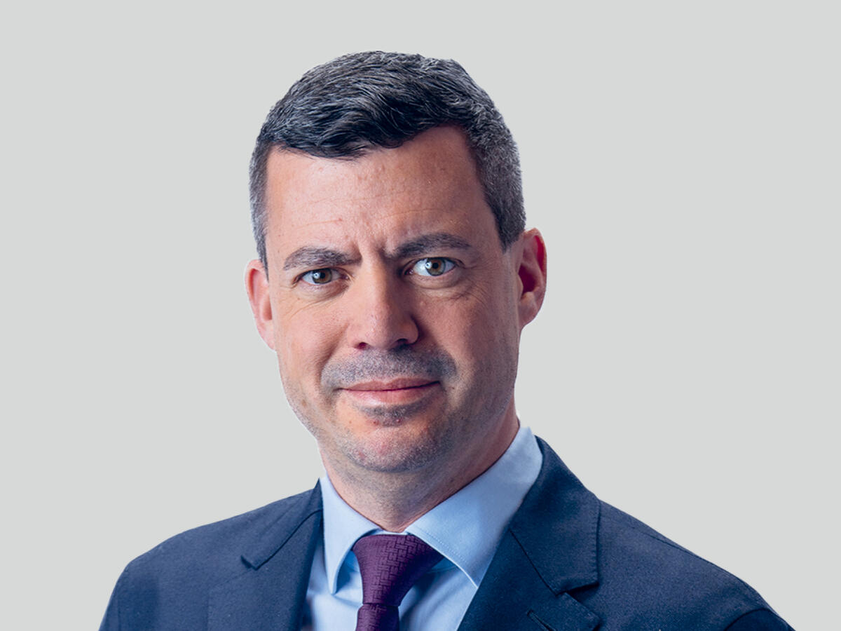 A professional portrait of a middle-aged man with short, dark gray hair, wearing a navy blue suit, a light blue dress shirt, and a dark purple tie. He has a confident and composed expression with a slight smile. The background is a neutral light gray gradient.