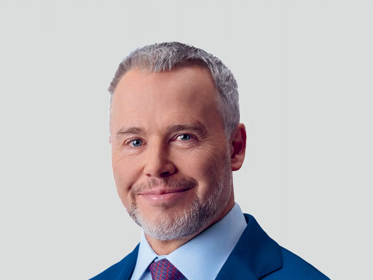 A professional portrait of a middle-aged man with short, neatly styled gray hair and a well-groomed beard, wearing a navy blue suit, a light blue dress shirt, and a red patterned tie. He has a confident and approachable expression with a slight smile. The background is a neutral light gray gradient.
