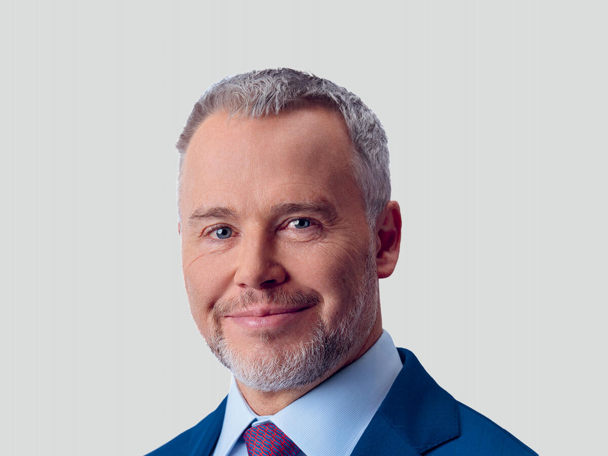 A professional portrait of a middle-aged man with short, neatly styled gray hair and a well-groomed beard, wearing a navy blue suit, a light blue dress shirt, and a red patterned tie. He has a confident and approachable expression with a slight smile. The background is a neutral light gray gradient.