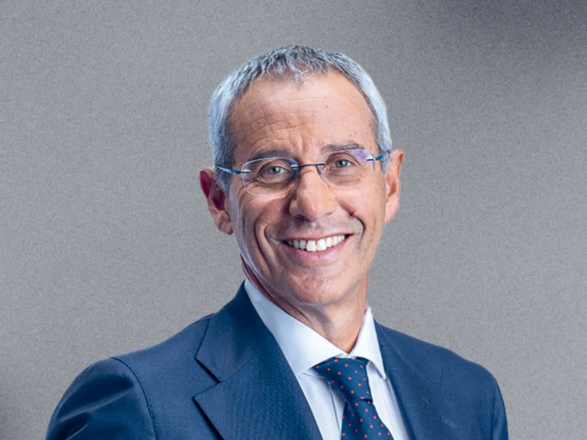 A professional portrait of a middle-aged man with short gray hair, wearing thin-framed glasses, a navy blue suit, a white dress shirt, and a dark blue tie with a subtle pattern. He has a warm and confident smile. The background is a neutral gray gradient.