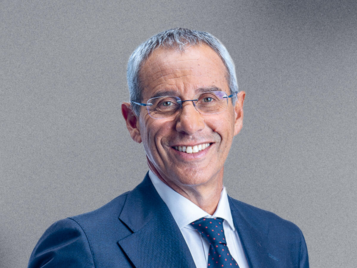 A professional portrait of a middle-aged man with short gray hair, wearing thin-framed glasses, a navy blue suit, a white dress shirt, and a dark blue tie with a subtle pattern. He has a warm and confident smile. The background is a neutral gray gradient.