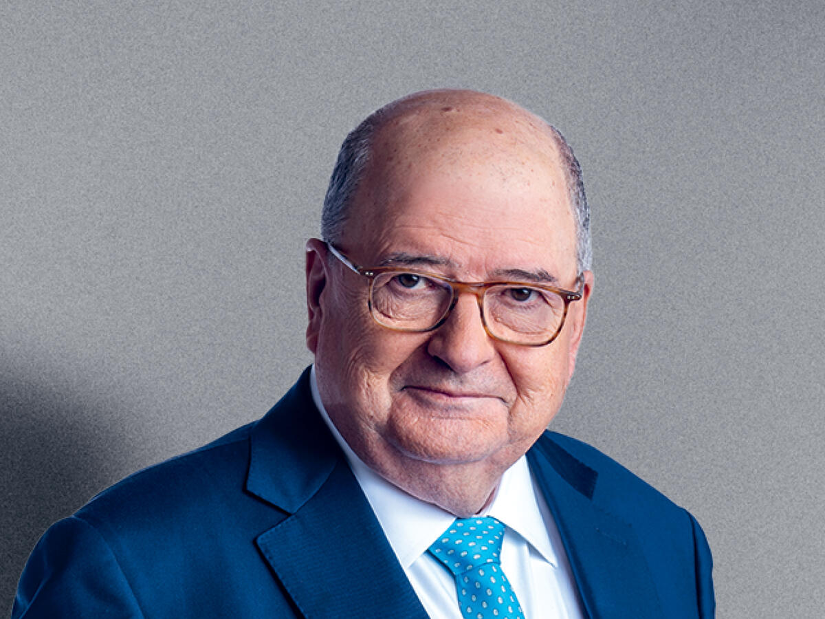 A professional portrait of an older man with a bald head and short gray hair on the sides, wearing brown-framed glasses, a navy blue suit, a white dress shirt, and a light blue tie with a subtle pattern. He has a calm and composed expression with a slight smile. The background is a neutral gray gradient.