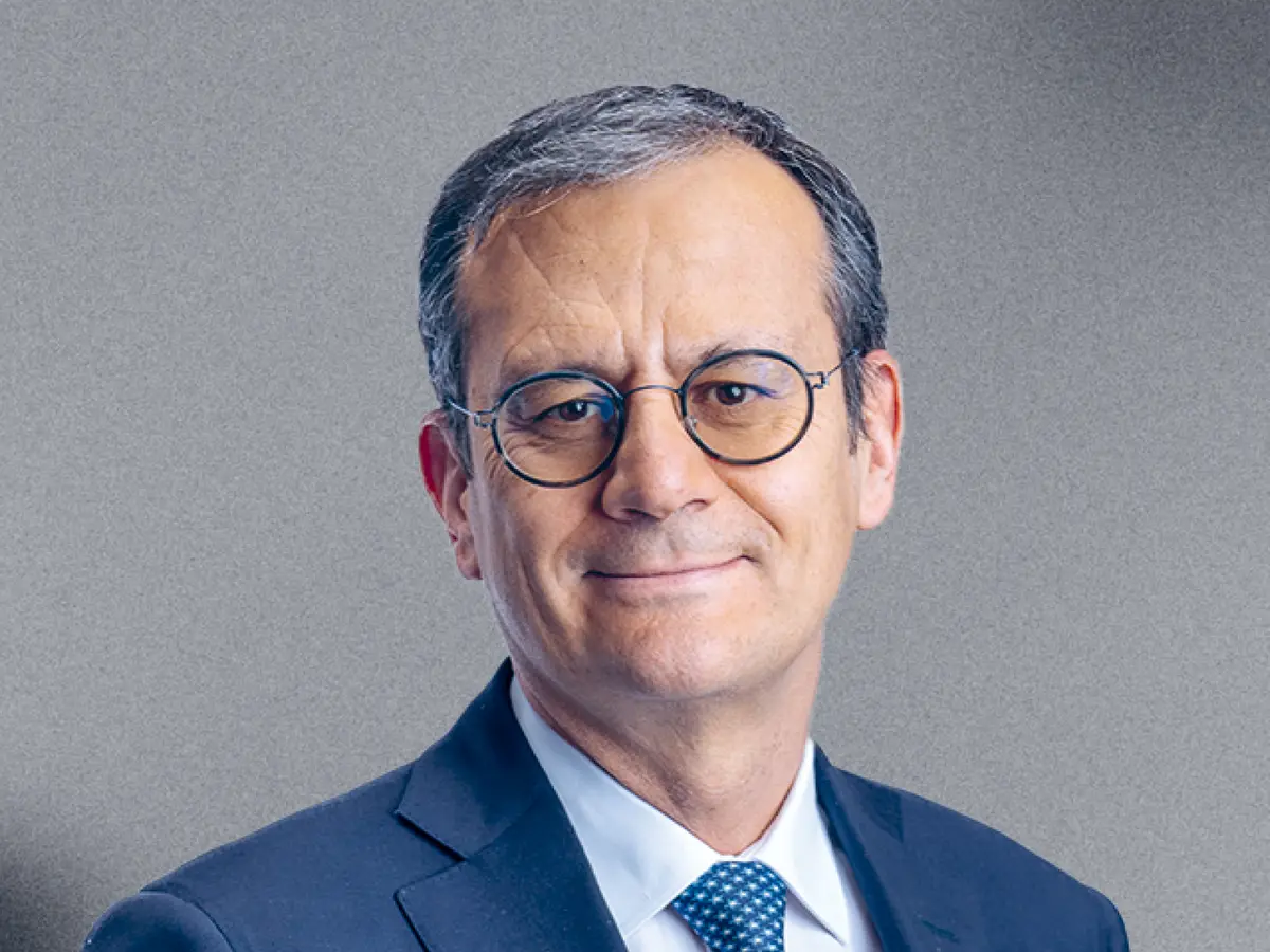 A professional portrait of a middle-aged man with short graying hair, wearing round glasses, a navy blue suit, a white dress shirt, and a blue patterned tie. He has a confident and approachable expression with a slight smile. The background is a neutral gray gradient.