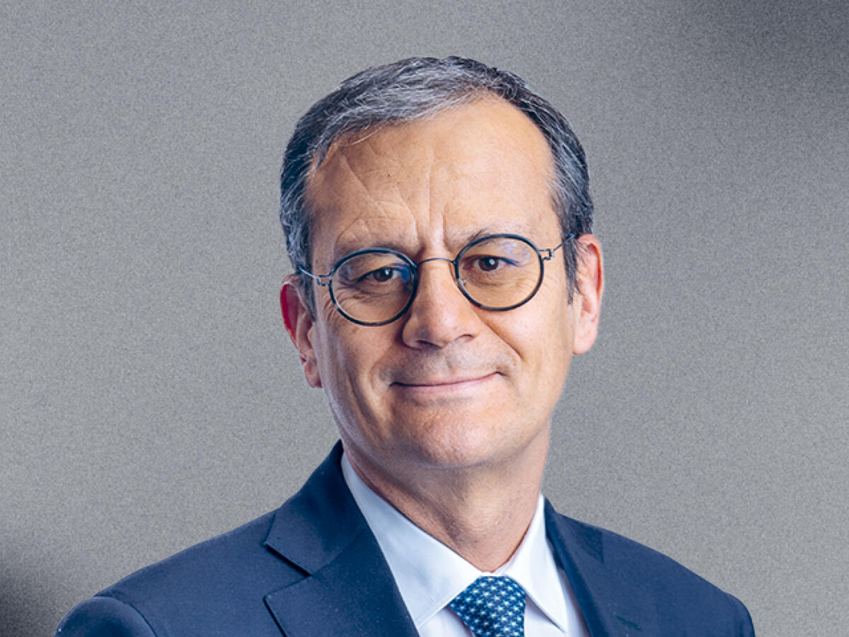 A professional portrait of a middle-aged man with short graying hair, wearing round glasses, a navy blue suit, a white dress shirt, and a blue patterned tie. He has a confident and approachable expression with a slight smile. The background is a neutral gray gradient.
