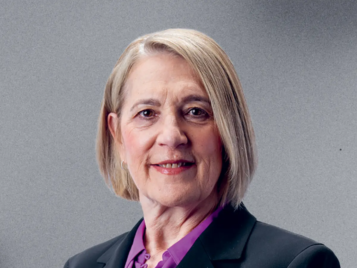 A professional portrait of a middle-aged woman with short, straight blonde hair, wearing a black blazer over a purple blouse. She has a warm and confident expression with a slight smile. The background is a neutral gray gradient.