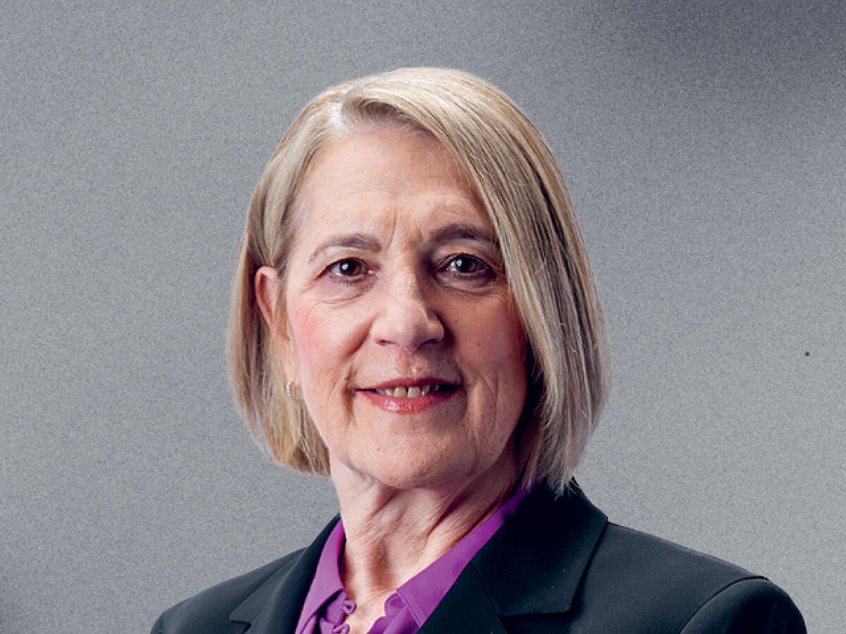 A professional portrait of a middle-aged woman with short, straight blonde hair, wearing a black blazer over a purple blouse. She has a warm and confident expression with a slight smile. The background is a neutral gray gradient.