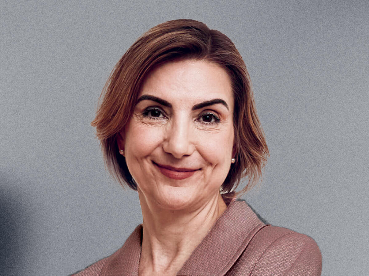 A professional portrait of a middle-aged woman with short, wavy brown hair, wearing a mauve-colored blazer with a subtle texture. She has a warm and confident expression with a gentle smile. The background is a neutral gray gradient.