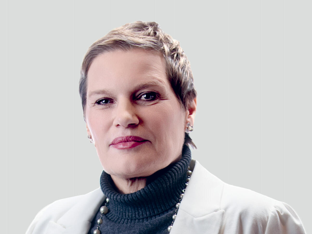A professional portrait of a middle-aged woman with short, styled blonde hair, wearing a white blazer over a dark turtleneck sweater. She is accessorized with pearl earrings and a matching pearl necklace. She has a confident and composed expression with a slight smile. The background is a neutral light gray gradient.