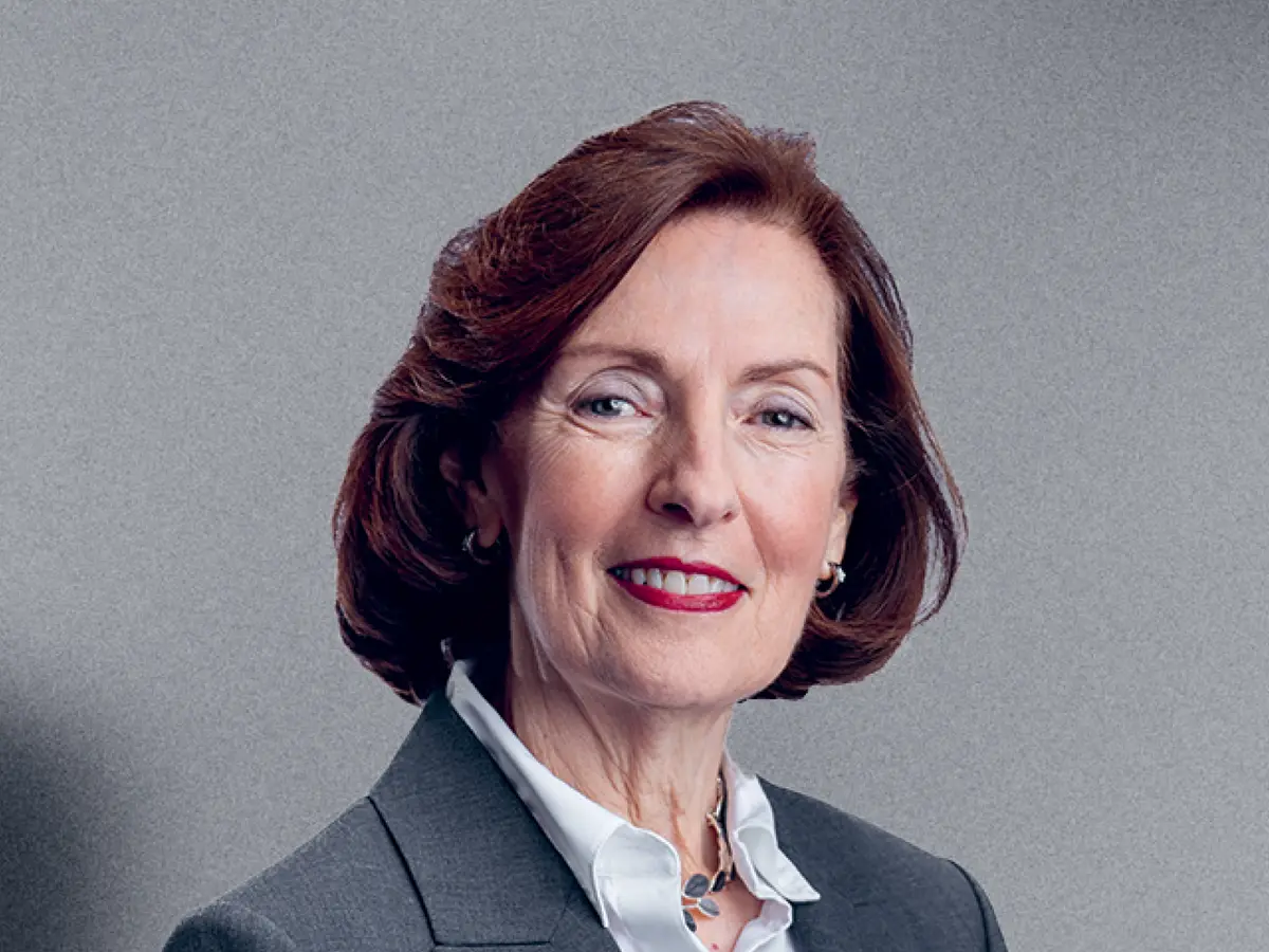 A professional portrait of a middle-aged woman with shoulder-length, wavy auburn hair, wearing a dark gray blazer over a white blouse. She is accessorized with a silver necklace and matching earrings. She has a confident and warm expression with a slight smile. The background is a neutral gray gradient.