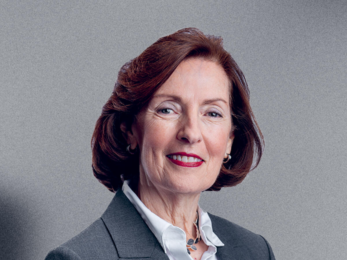 A professional portrait of a middle-aged woman with shoulder-length, wavy auburn hair, wearing a dark gray blazer over a white blouse. She is accessorized with a silver necklace and matching earrings. She has a confident and warm expression with a slight smile. The background is a neutral gray gradient.