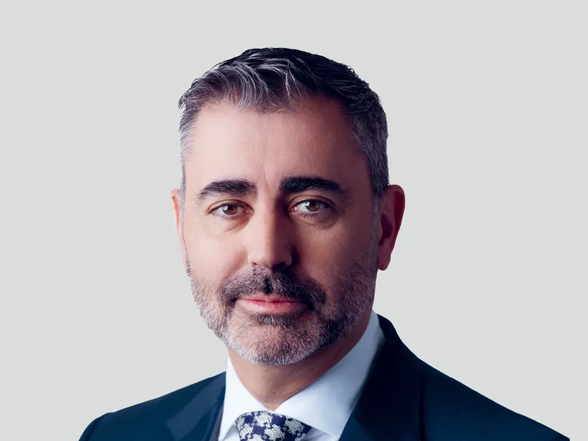 A professional portrait of a middle-aged man with short, dark gray hair and a well-groomed beard, wearing a dark navy suit, a white dress shirt, and a patterned tie. He has a confident and composed expression with a slight smile. The background is a neutral light gray gradient.
