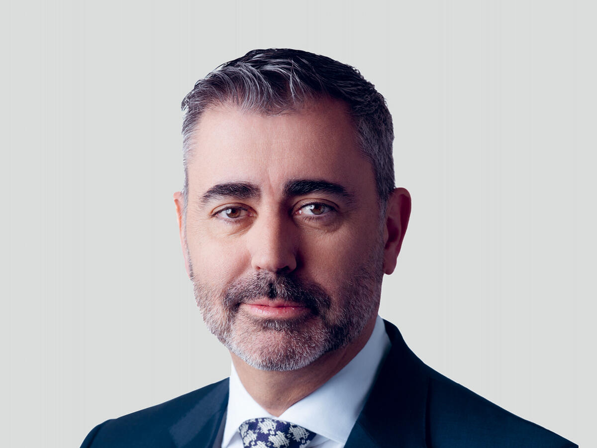 A professional portrait of a middle-aged man with short, dark gray hair and a well-groomed beard, wearing a dark navy suit, a white dress shirt, and a patterned tie. He has a confident and composed expression with a slight smile. The background is a neutral light gray gradient.