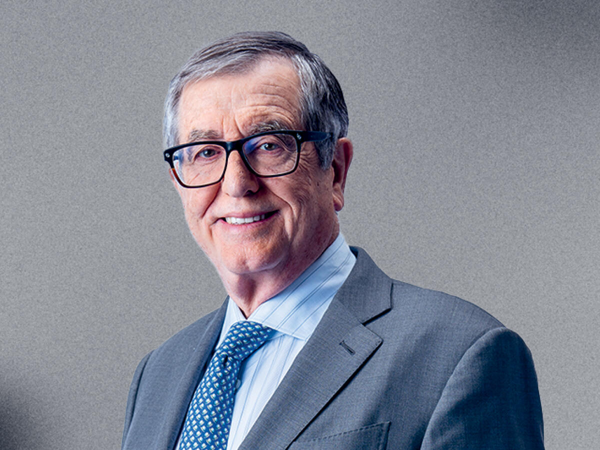 A professional portrait of an older man with short gray hair, wearing black-framed glasses, a gray suit, a light blue dress shirt, and a blue patterned tie. He has a friendly and confident expression with a slight smile. The background is a neutral gray gradient.