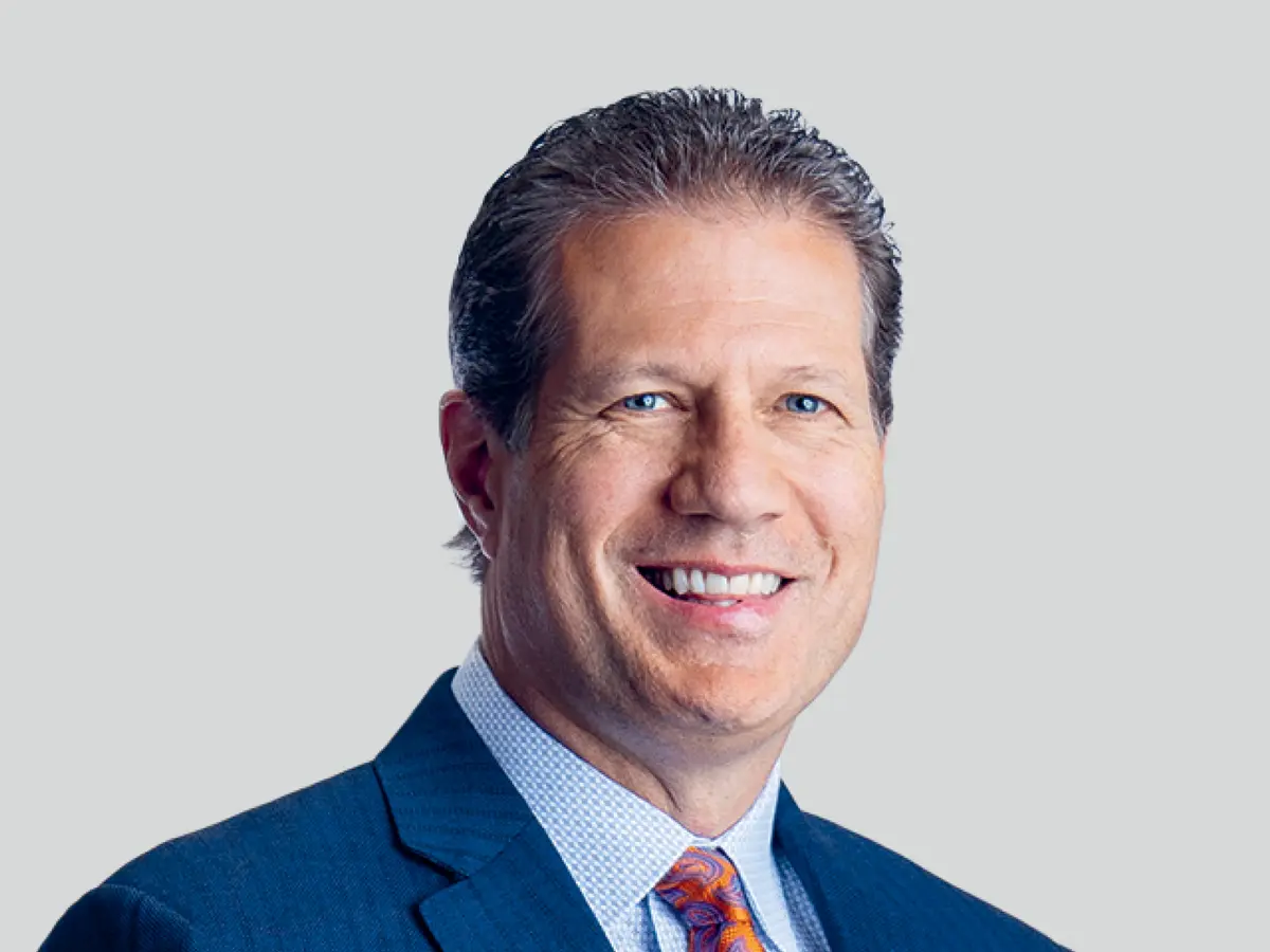 A professional portrait of a middle-aged man with short, wavy brown hair, wearing a navy blue suit, a light blue checkered dress shirt, and a patterned orange tie. He has a confident and friendly expression with a broad smile. The background is a neutral light gray gradient.