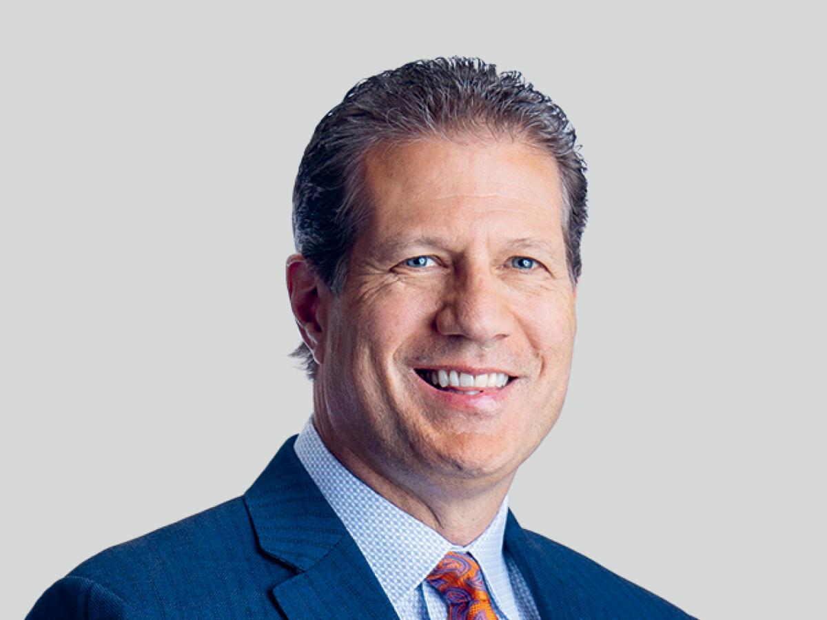 A professional portrait of a middle-aged man with short, wavy brown hair, wearing a navy blue suit, a light blue checkered dress shirt, and a patterned orange tie. He has a confident and friendly expression with a broad smile. The background is a neutral light gray gradient.
