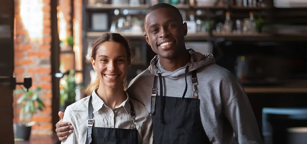A Black male employee is hugging a female coworker inside a store. Both are wearing aprons.